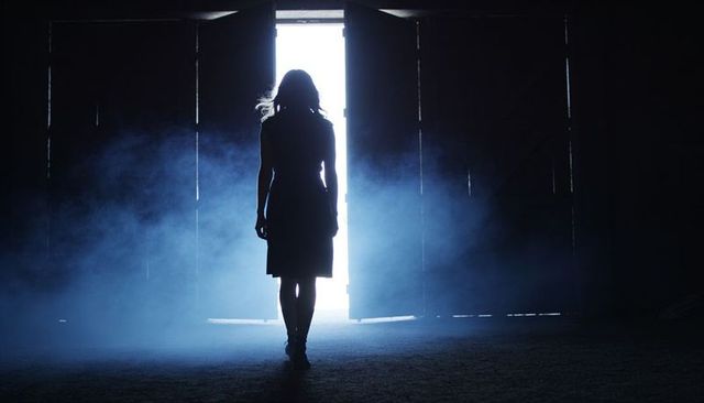 Woman walking through foggy barn doorway backlit by bright light, dramatic cinematic silhouette