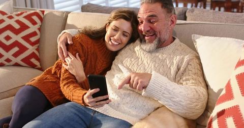 Couple cuddling on sofa while scrolling smartphone, cozy sweaters and warm living room