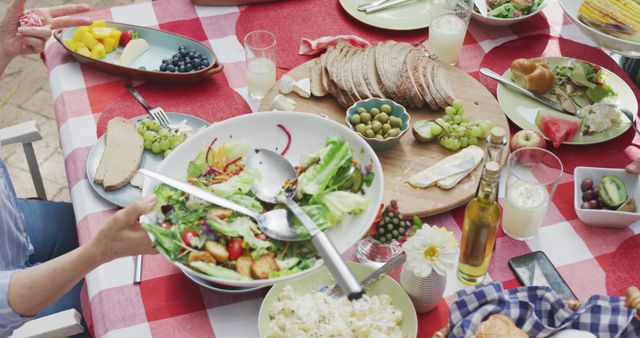 This close-up captures a group of friends enjoying a meal at a summer picnic table. The table features fresh salad, bread, fruit, and beverages, creating a vibrant and festive atmosphere. Ideal for promoting outdoor dining, summer gatherings, recipes, healthy eating, and social events.