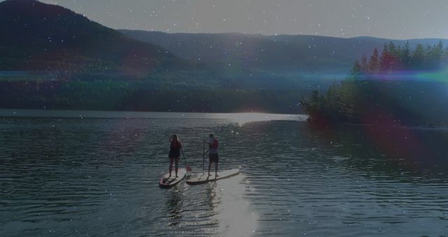 Couple engaging in paddleboarding on a calm lake during twilight. The peaceful natural setting is highlighted by clear skies and gentle water reflections, creating a serene and romantic atmosphere. Perfect for promoting outdoor recreational activities, travel destinations, adventure vacations, or romantic getaways.