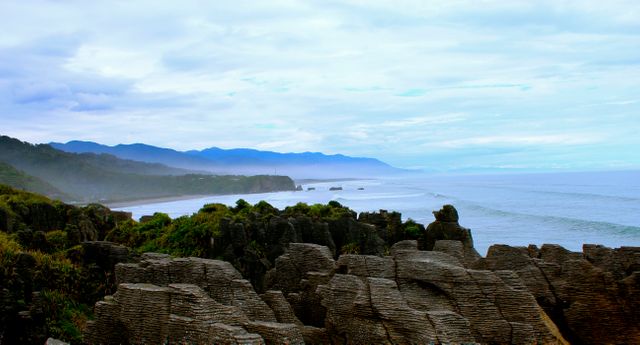 This image captures a picturesque coastal landscape featuring a rugged rocky shoreline with waves crashing against it. The background showcases a stunning view of mountains meeting the sea under a cloudy sky. Perfect for use in nature and travel blogs, environmental presentations, and tourism brochures promoting coastal destinations.