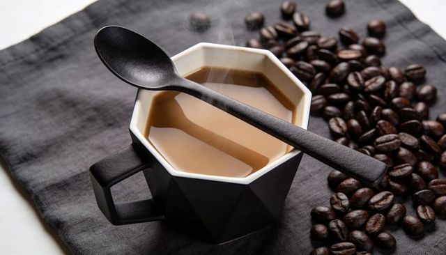 Steaming octagonal matte black mug with milky coffee, spoon and scattered coffee beans