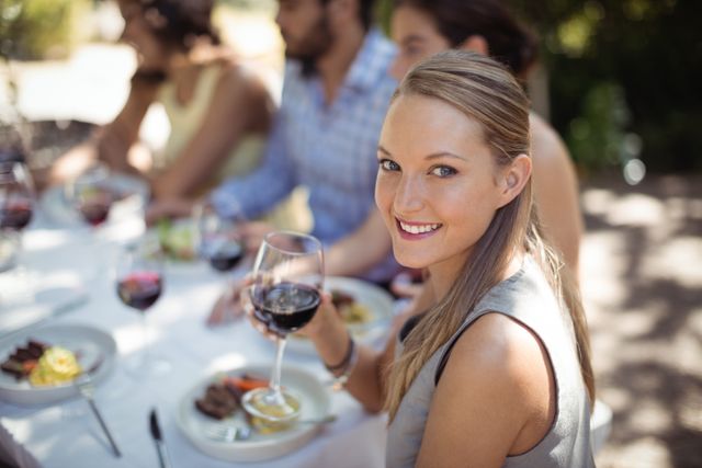 Woman holding a glass of wine while dining with friends at an outdoor restaurant. Ideal for use in lifestyle blogs, social media posts about dining out, advertisements for restaurants or wine brands, and articles on social gatherings and leisure activities.
