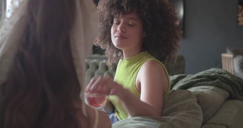 Diverse friends sharing tea on green sofa, relaxing with curly hair and cozy home vibes