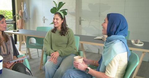 Diverse women sharing relaxed coworking conversation around table holding tablet and coffee
