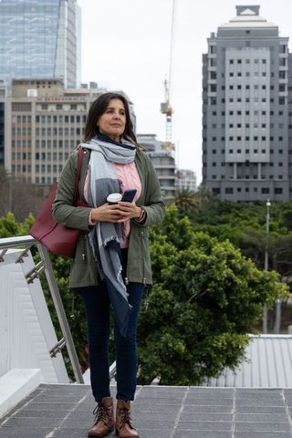 Middle-aged woman standing on urban rooftop holding coffee and checking smartphone