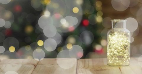 Glass jar glowing with warm fairy lights on wooden table with holiday bokeh, cozy festive ambience