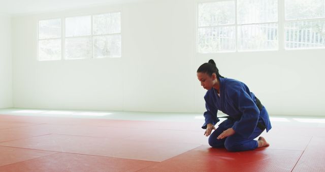This image shows a female judoka bowing on a red tatami mat inside an empty training room, emphasizing respect and discipline in martial arts culture. The natural light flowing through the large windows creates a serene setting, ideal for educational purposes in martial arts training materials, dojo promotions, or motivational posters related to discipline and focus in sports.