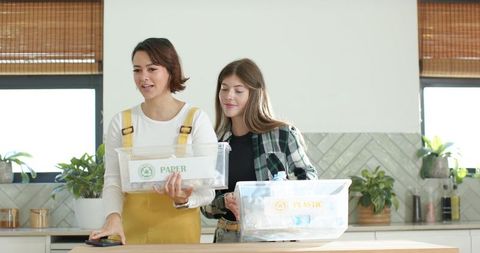 Two women sorting recycling at kitchen island holding paper and plastic bins, ecofriendly home