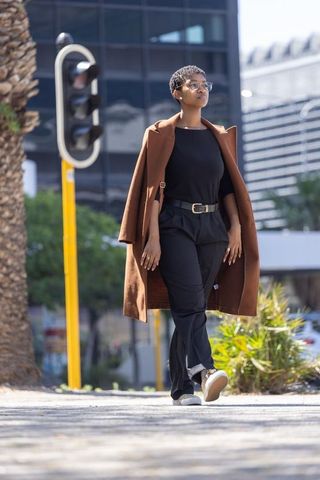 Confident african woman walking downtown wearing brown coat draped over shoulders and glasses