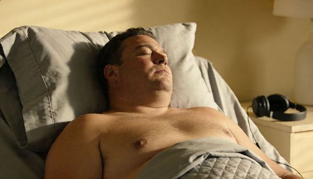 Shirtless middle-aged man resting in warm morning light with headphones on nightstand