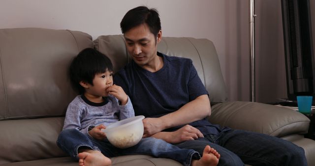 Father and young son sharing a snack while sitting on a sofa in the living room. Captures a tender moment of family bonding. Ideal for use in contexts of family life, parenting advice, home living, and lifestyle blogs.