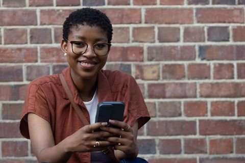 Smiling african american woman holding smartphone against red brick wall urban portrait