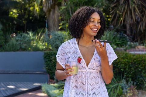 Smiling african american woman speaking into smartphone holding lemonade with strawberry on sunny pa