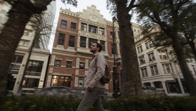 Young man walking past historic brick building carrying backpack on city sidewalk
