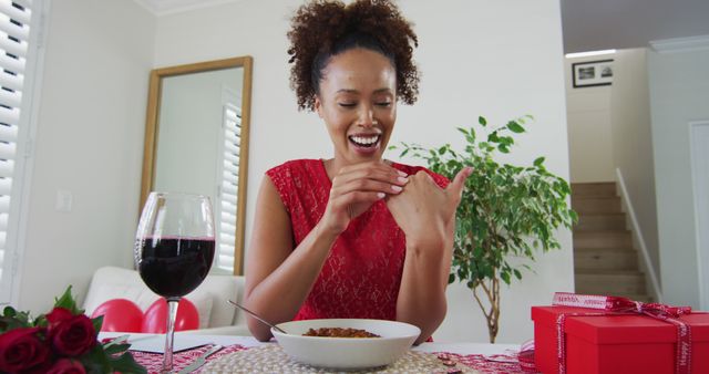 Excited woman wearing red dress displaying engagement ring during romantic dinner at home. Featuring wine glass, bouquet of roses, and gift boxes on table. Perfect for advertisements related to engagements, relationships, Valentine's Day, romantic celebrations, or lifestyle blogs focusing on special moments and love.