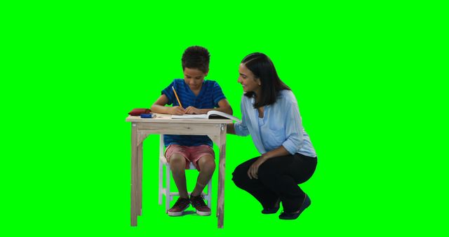 A young boy is focused on writing in a book at a desk, with a woman, a teacher, assisting him, with copy space. They are set against a green screen background, which allows for easy editing and insertion of different backgrounds in post-production.