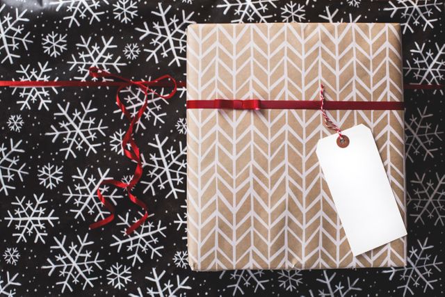 Present wrapped in beige and white patterned paper with a red ribbon placed on top of black snowflake wrapping paper. White blank gift tag attached. Ideal for holiday gift guide publications, Christmas blog posts, or holiday decoration ideas.