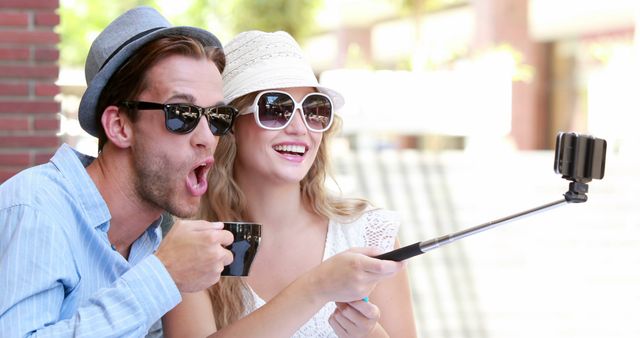 A young Caucasian couple enjoys taking a selfie with a smartphone on a selfie stick, with copy space. Their playful expressions and casual summer attire suggest a light-hearted moment during a leisurely day out.