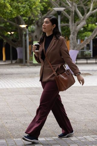 Young professional walking across campus plaza holding to-go coffee, wearing brown blazer