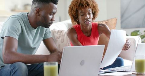 African american couple working from home reviewing documents with laptops on sofa