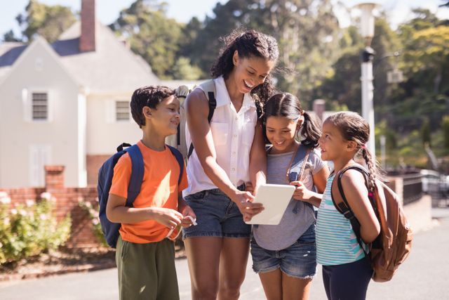Teacher and children are gathered around a digital tablet on a sunny street during a summer trip. They are smiling and appear engaged with the device. This image can be used for educational content, summer camp promotions, technology in education, and outdoor learning activities.