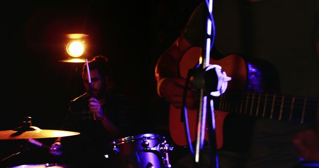 Musicians perform on stage, with the focus on a drummer and a guitarist, both appearing to be young adults. Low lighting sets an intimate atmosphere for a live music event, capturing the essence of a small venue concert.