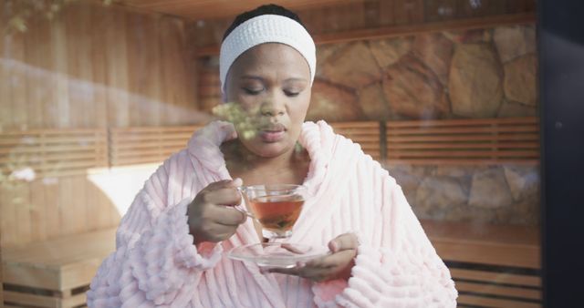 Woman in spa sipping herbal tea wearing pink robe, enjoying a calm and peaceful environment. Ideal for use in health and wellness promotions, spa advertisements, or articles on relaxation and self-care.