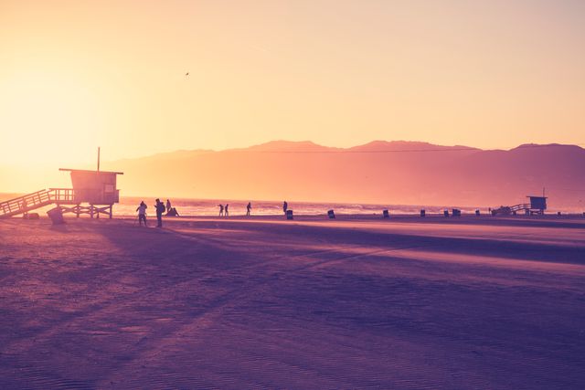 Beautiful scene of sunset casting a golden hue on the beach with silhouetted figures walking near the ocean and a lifeguard tower. Ideal for travel brochures, websites promoting beach destinations, or relaxation-themed advertising.