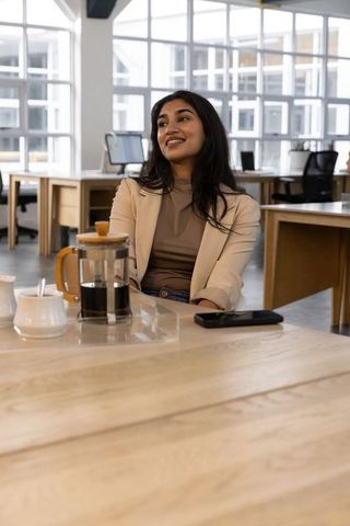 Indian professional wearing blazer sipping french press coffee at modern coworking desk, smiling