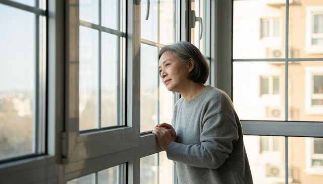Senior woman leaning and gazing through window in urban apartment, contemplative morning light