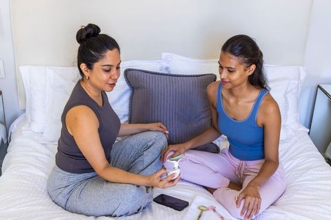 Two women sharing handheld beauty device and chatting on cozy bed during at-home skincare routine