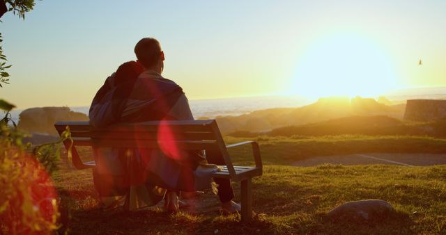A young Caucasian couple enjoys a serene sunset together, sitting on a bench overlooking a scenic landscape, with copy space. Their shared blanket and the warm glow of the setting sun create a romantic and peaceful atmosphere.