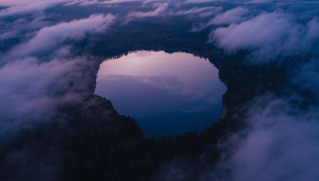 Aerial twilight view of circular mirrorlike lake reflecting sky amid foggy conifer forest