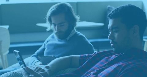 Two men scrolling smartphones and tablet on lounge couch, casual tech collaboration