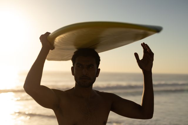 Young man holding a surfboard on his head while standing on the beach during sunset. Ideal for use in advertisements, travel blogs, fitness and lifestyle magazines, and promotional materials for surfing and beach-related activities.