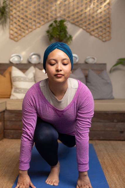Biracial woman in a hijab performing a stretching yoga pose on a blue mat in a cozy living room. This image exudes a sense of tranquility and mindfulness, ideal for promoting wellness, fitness, and inclusivity. Perfect for use in health and fitness blogs, social media, yoga classes, and advertisements focused on home exercise, relaxation, and well-being.
