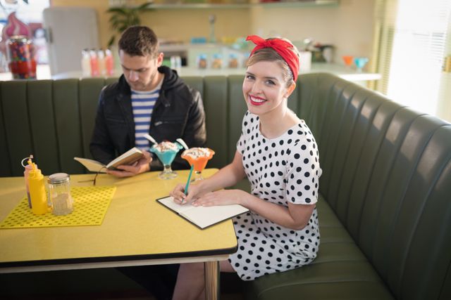 Young couple enjoying time in a retro diner. Woman in polka dot dress and red headband smiling while writing in a notebook, man reading a menu. Ice cream sundaes on the table. Ideal for themes of nostalgia, vintage fashion, classic American diners, and romantic dates.