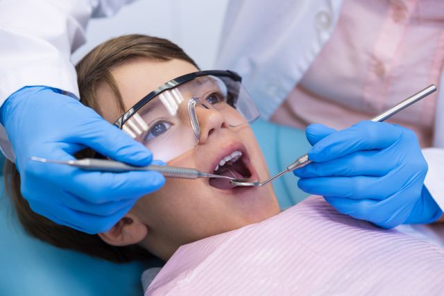 Young boy receiving a dental checkup at a clinic. The child is wearing protective eyewear and a dental bib while the dentist uses dental tools to examine his mouth. This image can be used for promoting dental health, pediatric dentistry services, or educational materials on oral hygiene for children.
