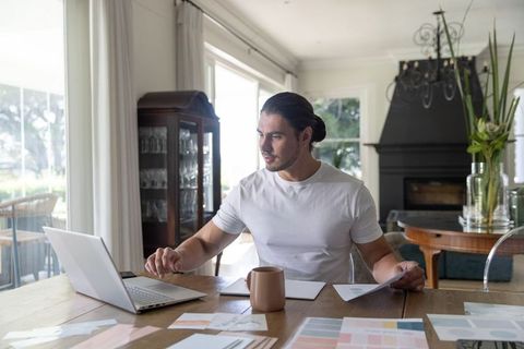 Young designer reviewing color swatches on laptop at sunlit dining table in modern home
