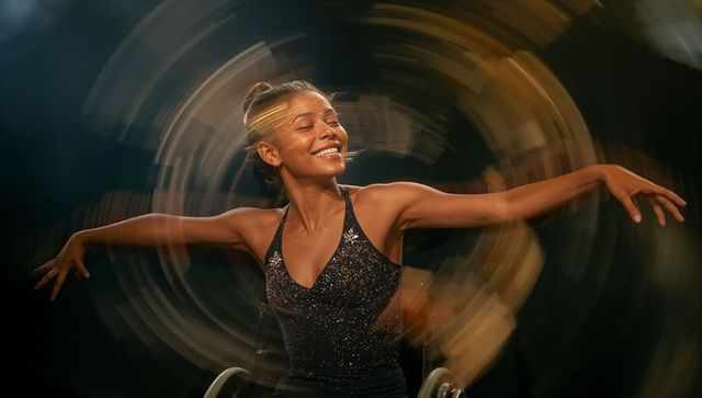 Spinning wheelchair dancer smiling with arms extending in sparkling halter dress on dark stage
