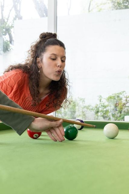 Young woman leaning over pool table aiming cue while partner holding bridge at home