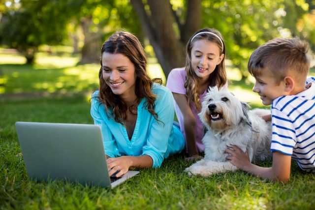 Family spending quality time together in a park, with mother using laptop and children playing with dog. Ideal for themes related to family bonding, outdoor activities, technology in nature, and leisure time.