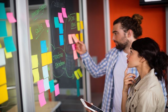 Male and female executives brainstorming ideas using colorful sticky notes on a glass wall in a modern office. This image can be used for business presentations, articles on teamwork and collaboration, project management materials, and creative strategy sessions.