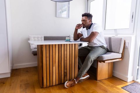 African american man sitting at modern wooden breakfast nook holding pen, contemplating
