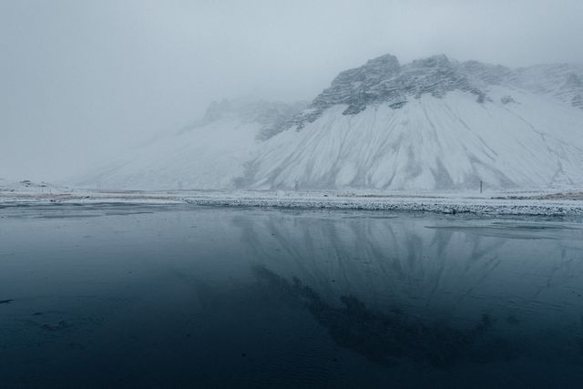 Snow-covered mountains reflect in an icy lake, creating a tranquil and dramatic winter landscape. This serene scene, with its overcast sky and cold weather, highlights the natural beauty of the area. Ideal for use in travel blogs, nature photography collections, winter tourism promotions, and scenic landscape advertisements.