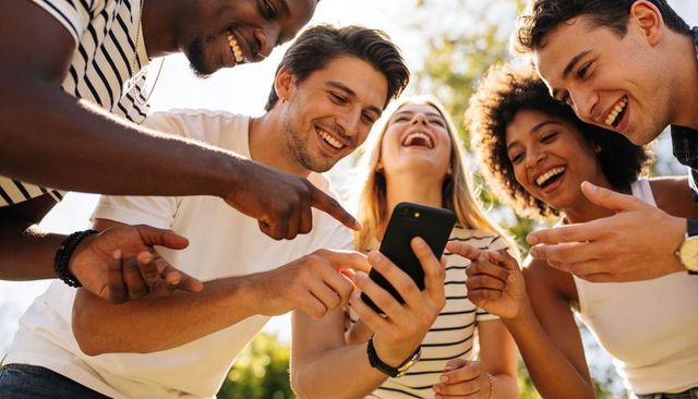 Young diverse group laughing, pointing and sharing smartphone in sunlit park