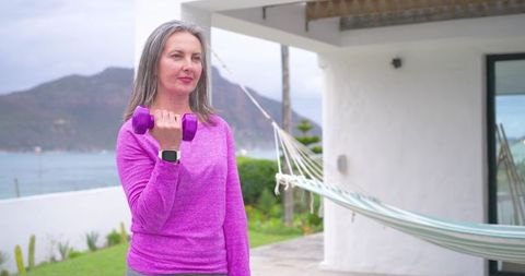 Mature woman exercising on sea terrace holding purple dumbbell wearing pink top smartwatch