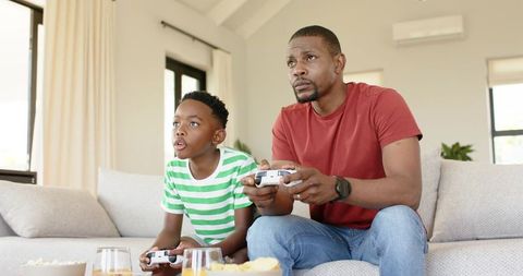 African American Dad and Son Playing Video Games on Couch Enjoying Family Bonding and Snacks