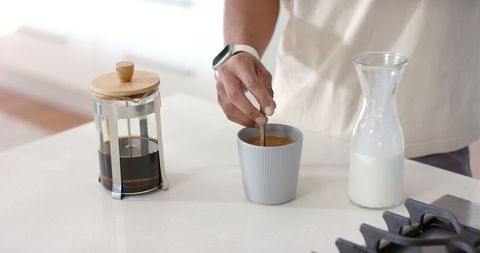 African american man stirring morning coffee at kitchen counter with french press and milk